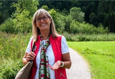 A friendly woman smiles on a hiking path surrounded by green nature. In the background, trees and a sunny landscape can be seen.