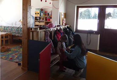 A bright room with wooden furniture and colorful play mats. A caregiver is helping a child get dressed.