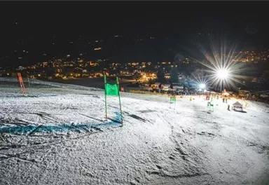 Eine beleuchtete Skipiste mit grünen Fahnen und Schnee. Im Hintergrund sind die Lichter einer Stadt bei Nacht zu sehen.