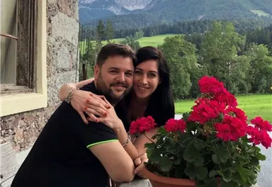 A couple sits happily at an outdoor table, embracing each other. In the foreground, there is a flowerpot with red geraniums, and in the background, green meadows and mountains can be seen.