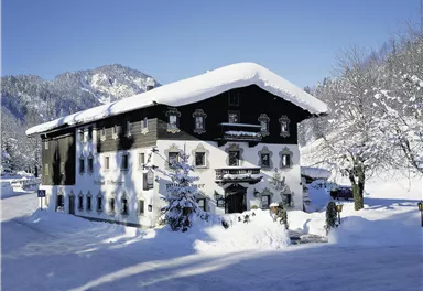 Ein traditionelles altes Haus im Schnee mit einem weißen Dach. Die Umgebung ist winterlich und malerisch, umgeben von Bergen und Bäumen.