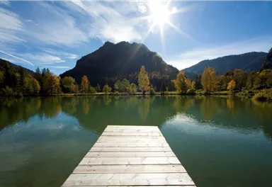 Ein ruhiger See mit einem Holzsteg und malerischen Bergen im Hintergrund. Die Herbstfarben der Bäume spiegeln sich im Wasser.