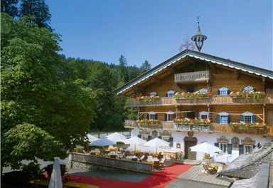 Ein traditionelles Alpenhaus mit blühenden Balkonen und einem roten Teppich vor dem Eingang. Umgeben von grünen Bäumen und einer einladenden Terrasse.
