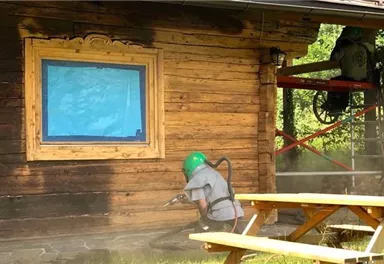 Ein Handwerker reinigt eine Holzwand mit einem Hochdruckgerät. Im Hintergrund ist ein Gerüst und ein blauer Fensterrahmen zu sehen.