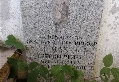 A commemorative plaque made of gravestone material, partially overgrown with plants. The text is difficult to read and is located on a wall.
