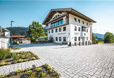 A modern building with a well-kept forecourt and blooming flowers. In the background, mountains and a clear sky are visible.