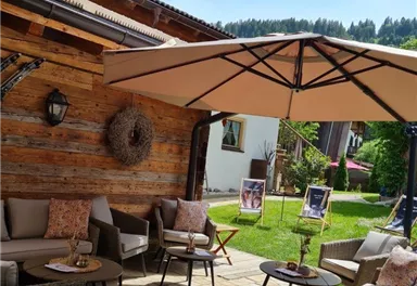 A cozy outdoor area with seating and a parasol. In the background, a well-maintained garden and a wooden house can be seen.