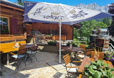A cozy terrace with wooden furniture and a large sun umbrella. In the background, green trees and mountains can be seen.
