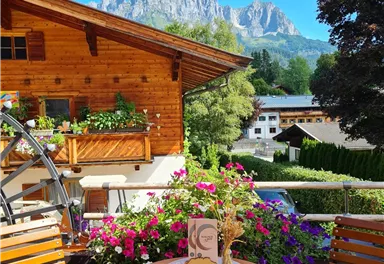 A cozy terrace with a round wooden table and chairs. In the background, there is a mountain panorama and colorful flowers.