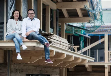 A couple is sitting relaxed on a construction scaffold. In the background, construction work on a modern building can be seen.