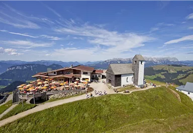 A picturesque mountain landscape with a cabin sign and a small church. Many people are enjoying the view on the terrace.