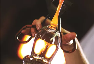 Ein Handwerker formt ein gläsernes Objekt mit Werkzeugen. Das Glas ist heiß und leuchtet orange in der Hintergrundbeleuchtung.