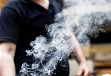 A glassblower works intently with hot glass. Smoke rises as he shapes the orange glass mass.