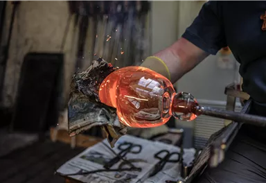 A glassmaker shapes a hot, glowing glass ball. Sparks fly as he works the glass.