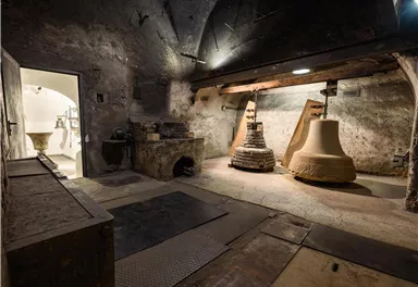 Ein historischer Glockenturm mit zwei großen Glocken. Der Raum hat eine rustikale, steinerne Atmosphäre und eine Tür, die zu einem weiteren Bereich führt.