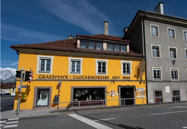 Ein gelbes Gebäude mit dem Schriftzug "Grassmayr - Glockengießerei seit 1599" steht an einer Straßenecke. Daneben befindet sich ein graues Haus und im Hintergrund sind Berge zu sehen.