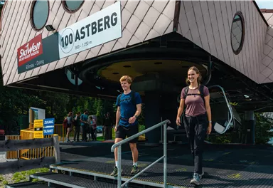 Two hikers are walking down a staircase from a modern mountain railway station. In the background, there are more people and a picturesque landscape visible.