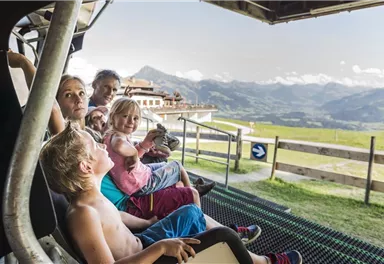 A family is sitting on a gondola outdoors, looking at the mountains. The sky is clear and the landscape is green and mountainous.
