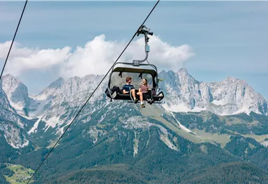 Zwei Personen in einem Sessellift vor dem Bergmassiv des Wilden Kaisers unter blauem Himmel.