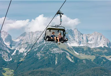 Two people are sitting in a gondola on their way over the mountains. The landscape consists of green forests and majestic peaks under a clear sky.