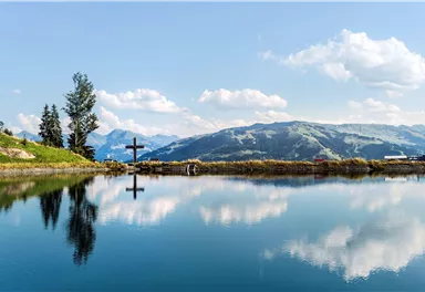 A quiet lake with clearly reflected mountains and a cross on the shore. The sky is blue with white clouds.