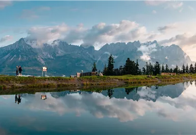 An impressive mountain landscape with mirrored water in the foreground. The sky is partly cloudy, and trees frame the scene.