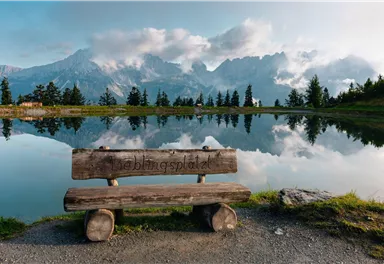 A wooden bench stands by the shore of a calm lake, surrounded by mountains. The sky is partly cloudy and reflects in the water.