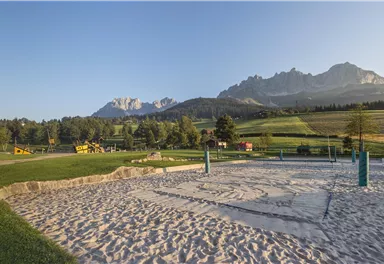 Ein Spielplatz mit Sandkasten und Blick auf die Berge. Die Umgebung ist grün und sonnig.