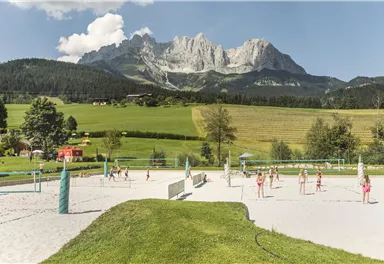 Ein Sandplatz mit Volleyballspielern vor beeindruckenden Bergen. Die Umgebung ist grün und sonnig, ideal für sportliche Aktivitäten.