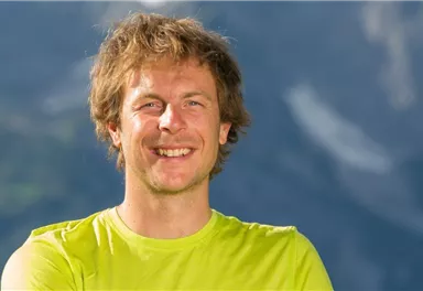 A smiling man is wearing a bright green shirt. Blurred mountains are visible in the background.