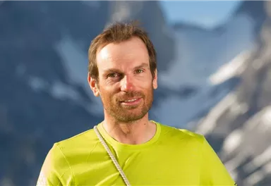 A man stands in front of a mountainous backdrop. He is wearing a light green sports shirt and has a friendly expression.