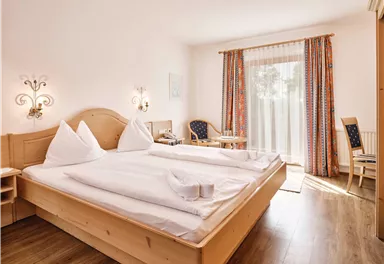 A cozy bedroom with a large bed and bright pillows. The room has a window with curtains and a small table with chairs.