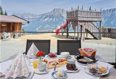 Ein gedeckter Tisch mit einem Frühstück im Freien, darunter Gebäck, Marmelade und Kaffee. Im Hintergrund ist ein Spielplatz mit einer Rutsche und den Bergen zu sehen.