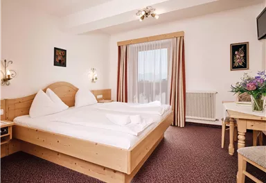 A cozy hotel room with a double bed and wooden furniture. The windows are furnished with curtains and a small flower vase is on the table.
