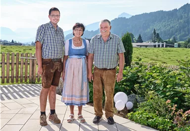 A group of six people in traditional costumes stands in front of an impressive mountain landscape. In the background, green forests and high mountains are visible.