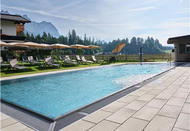 A beautiful pool with clear water, surrounded by green meadows and mountains. Loungers and sun umbrellas invite you to relax.