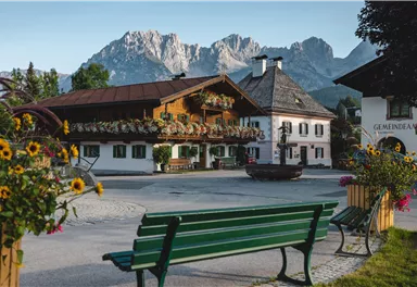 A picturesque village view with traditional Bavarian houses and impressive mountains in the background. Colorful flowers adorn the surroundings and a bench invites you to linger.