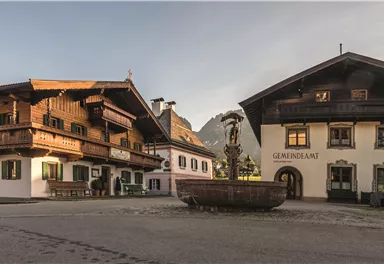 An idyllic village square with traditional wooden buildings and a fountain in the middle. The surroundings are quiet and surrounded by mountains.