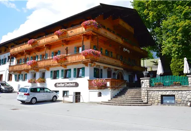 A traditional wooden house with balconies decorated with flowers. Additionally, a green surrounding and a parking lot in front.