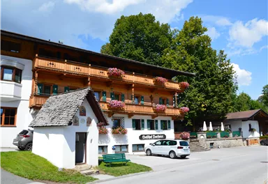A traditional old building with a wooden balcony and colorful flowers. It is located in a quiet environment with trees and an asphalt road.