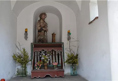 A small chapel with a statue and an altar. Flowers and candles decorate the space.