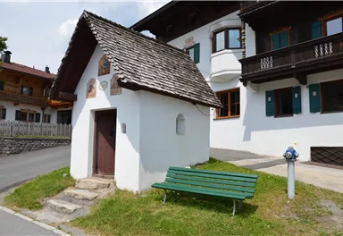 A small, white building with a sloping roof stands by the road. Next to the building is a green bench.