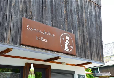 A wooden sign with the inscription "Eismanufaktur kElSer". In the background, a modern building with a clear sky can be seen.
