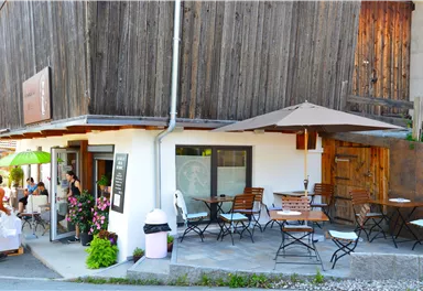 A cozy café with wooden paneling and outdoor tables. There are sun umbrellas and flowers in front of the entrance.