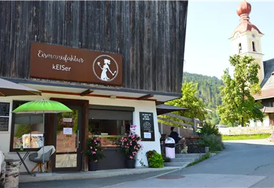 A small shop with a wooden facade and a sign with the name "Eismanufaktur kEISer". In the background, a church with a red tower and surrounding green landscape is visible.