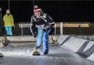A man is playing curling on an ice surface. In the background, two spectators in warm clothing can be seen.