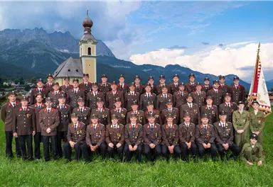 A group of men and women in uniform stands in a green meadow in front of a church with mountains in the background. They are posing together for a group photo.