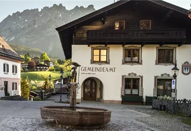Ein malerisches Dorf mit traditionellen Häusern und einem schönen Brunnen. Im Hintergrund sind beeindruckende Berge und eine grüne Landschaft zu sehen.