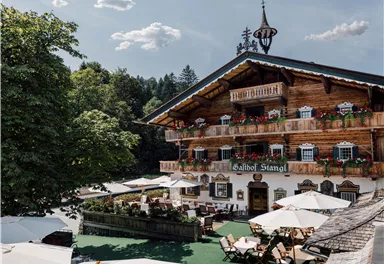 Ein traditionelles Alpenhaus mit blühenden Balkonen und einem roten Teppich vor dem Eingang. Umgeben von grünen Bäumen und einer einladenden Terrasse.
