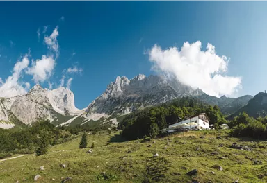 Eine malerische Berglandschaft mit hohen Felsen und frischem Grün. Im Vordergrund steht ein einfaches Gebäude, umgeben von Natur.